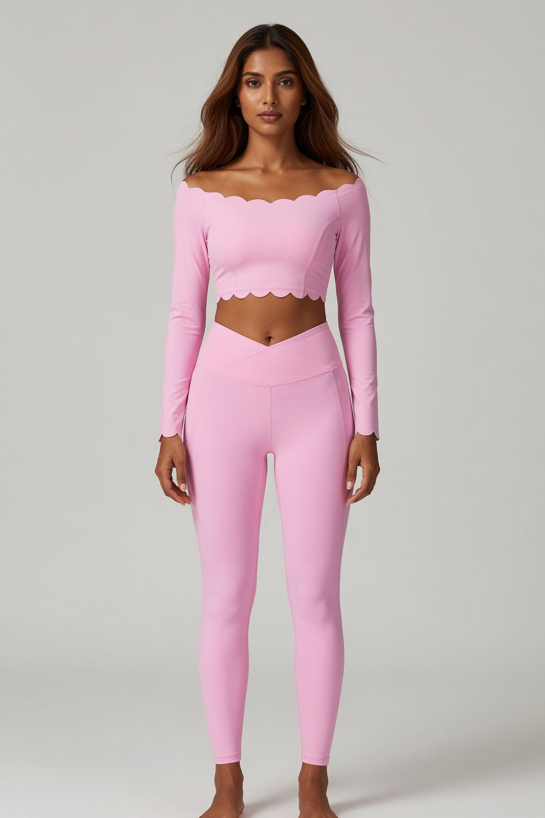 A woman modeling a light pink two-piece activewear set consisting of a long-sleeve cropped top with a scalloped neckline and matching high-waisted leggings, shot against a neutral light gray background.