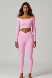 A woman modeling a light pink two-piece activewear set consisting of a long-sleeve cropped top with a scalloped neckline and matching high-waisted leggings, shot against a neutral light gray background.