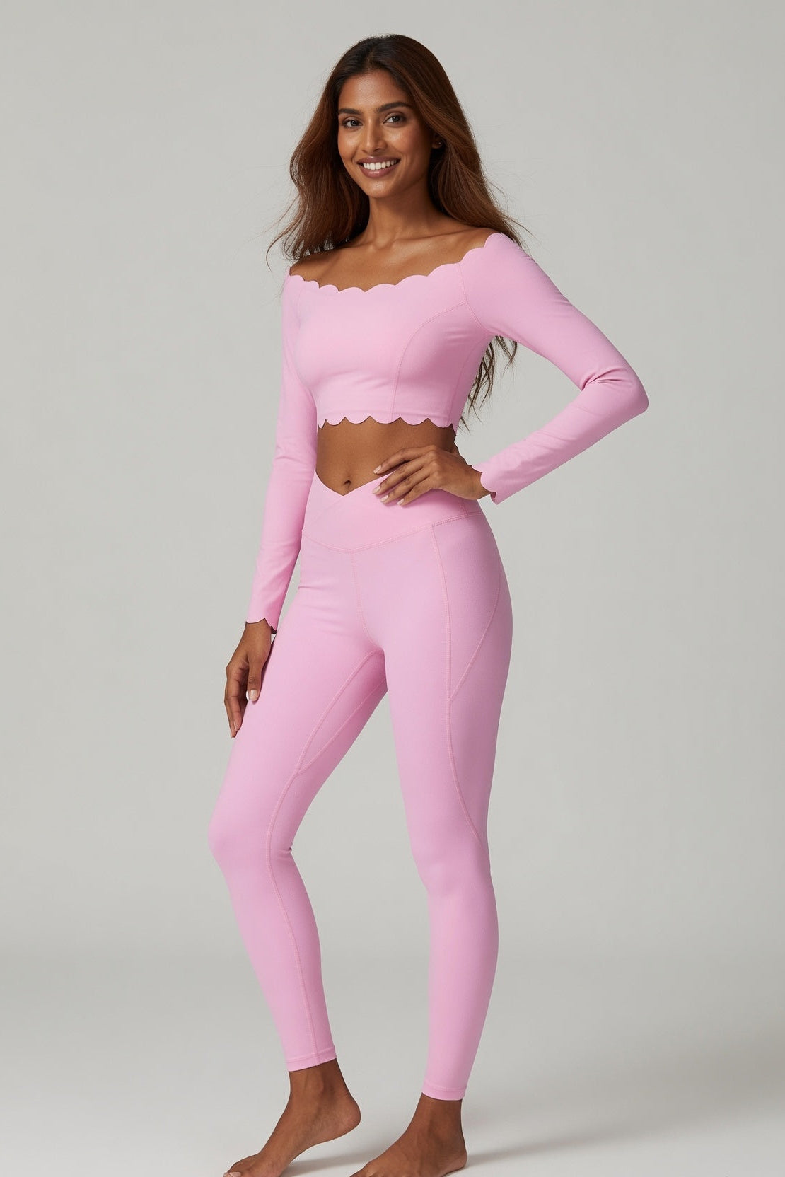A woman modeling a light pink two-piece activewear set consisting of a long-sleeve crop top with a scalloped neckline and matching high-waisted leggings, photographed against a neutral light gray studio background.