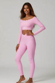 A woman modeling a light pink two-piece activewear set consisting of a long-sleeve crop top with a scalloped neckline and matching high-waisted leggings, photographed against a neutral light gray studio background.